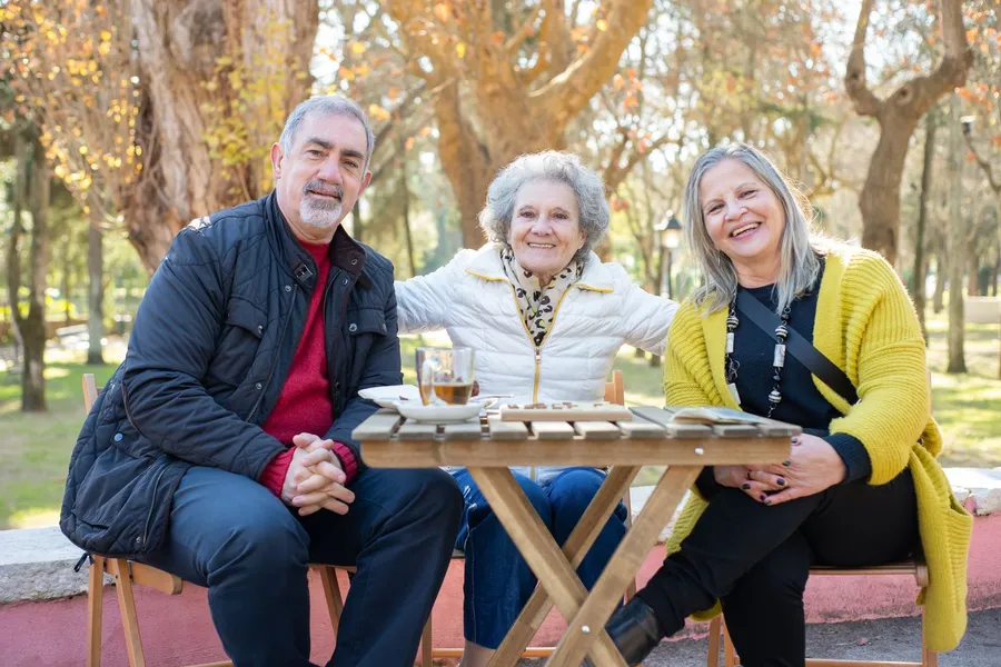 Three older adults smile at a wooden table in a park on a sunny autumn day