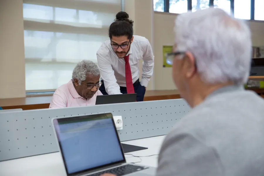 An older man in a pink shirt works on a laptop while a younger volunteer leans in to help, in a bright computer room