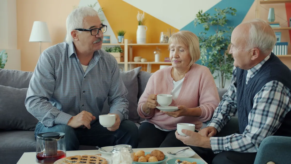Three older adults sit on a gray sofa drinking tea and talking, with cookies and a pie on the coffee table
