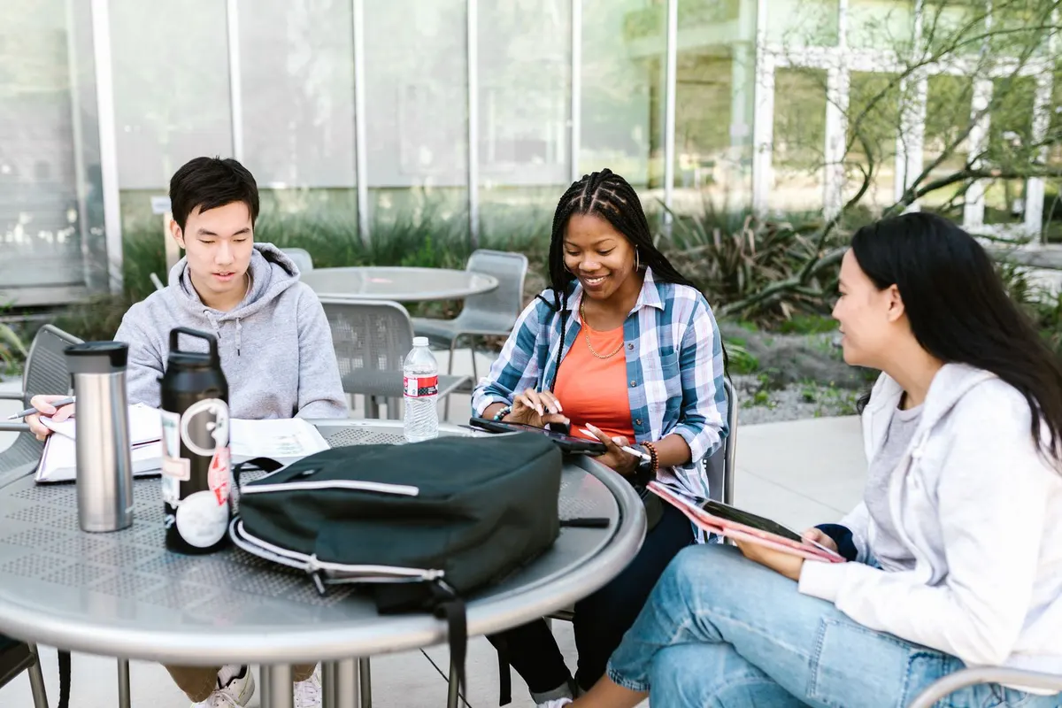 Tres estudiantes universitarios sentados en una mesa redonda al aire libre en el campus, sonriendo y trabajando con cuadernos y tabletas