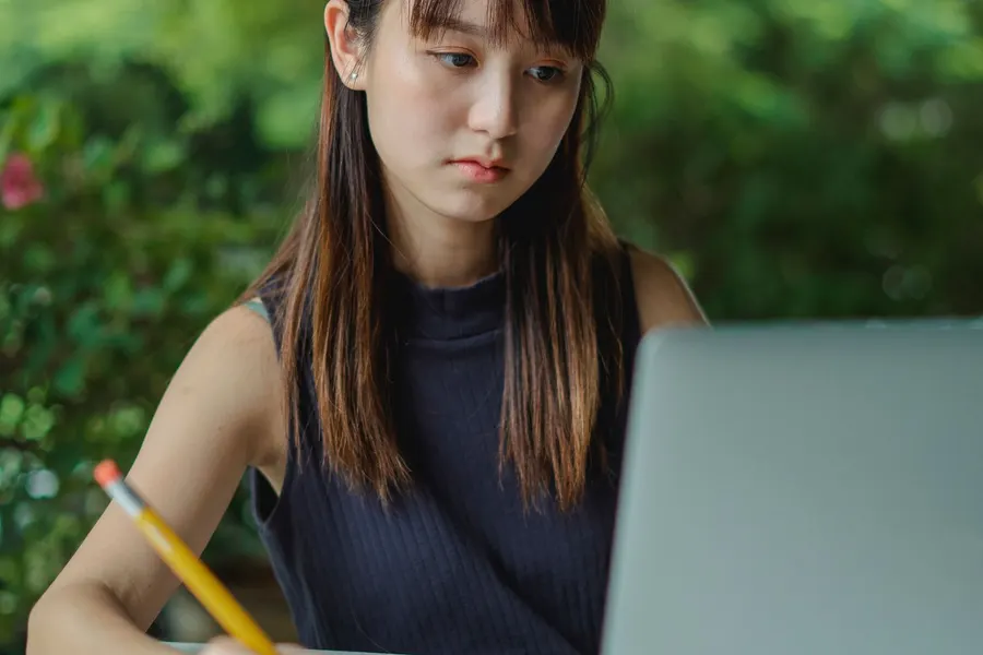 Una joven sentada en una mesa al aire libre escribe con un lápiz en un cuaderno mientras mira su computadora portátil