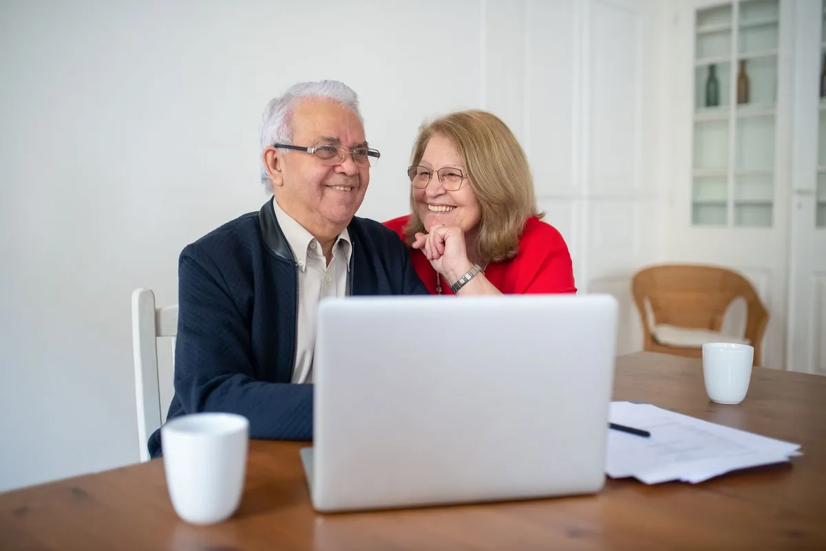 An older couple sitting at a dining table, smiling while looking at a laptop with papers and coffee cups beside them