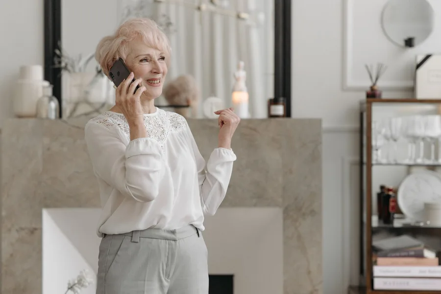An older woman standing in her living room, smiling while talking on a mobile phone