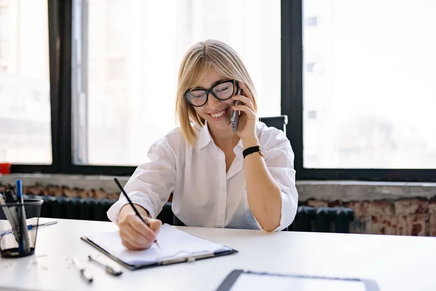 A smiling woman in glasses talks on the phone and writes notes on a clipboard at a desk by a window
