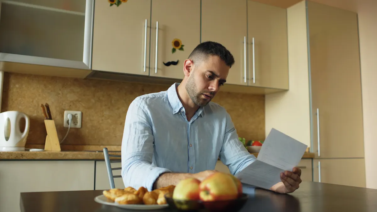 A man in a light blue shirt sits at a kitchen table reading a letter, with a bowl of apples in front of him