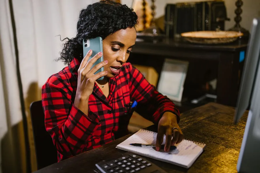 A woman on a phone call takes notes in a notebook at a wooden desk