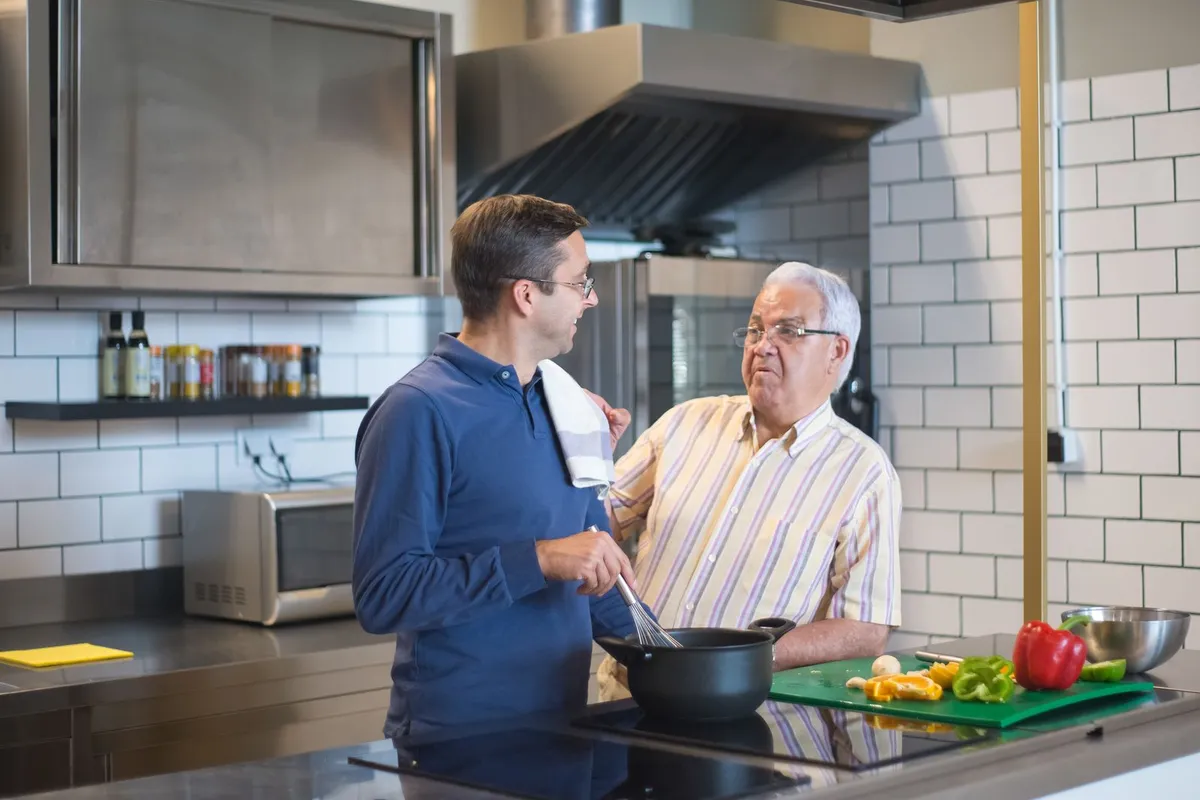 A middle-aged man in a blue shirt stirs a pot while an older man in a striped shirt talks with him in a kitchen