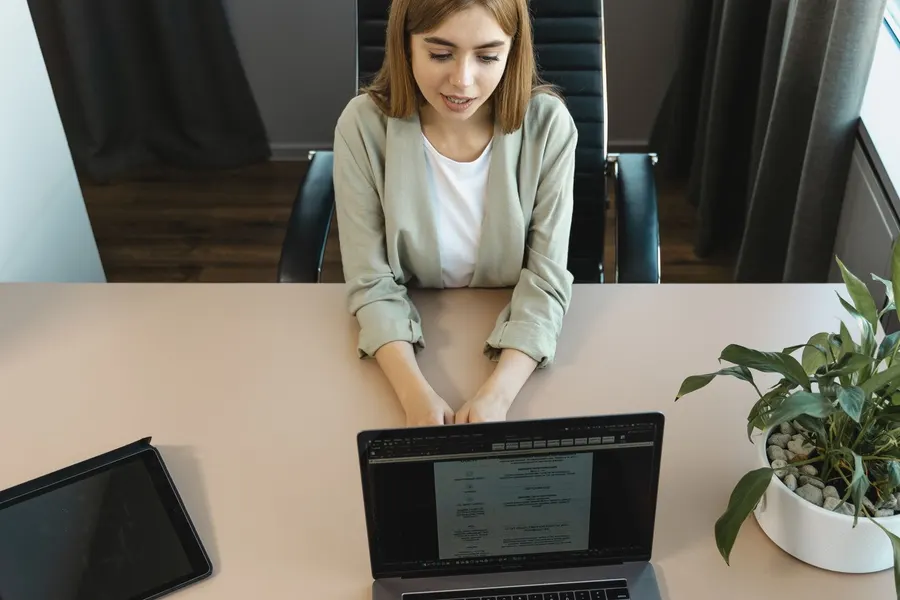 A woman sitting across a desk from an interviewer who is reading her paperwork, with a laptop and a plant on the table