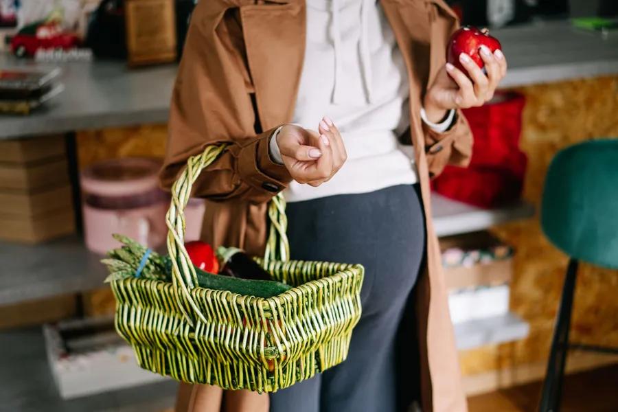 A pregnant woman in a tan coat holds a woven basket with vegetables and an apple