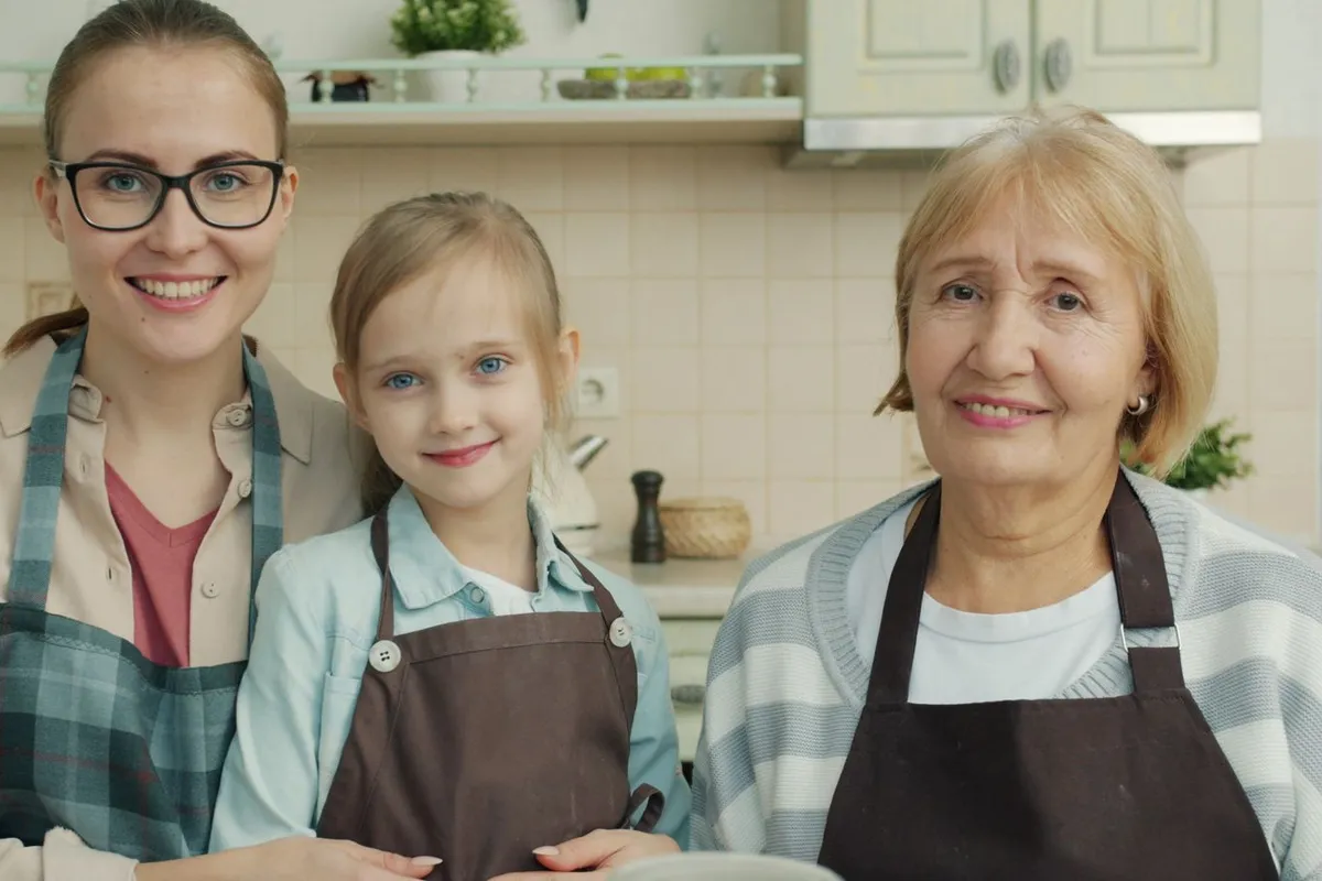 A smiling young woman, a little girl and an older woman in aprons stand together in a home kitchen