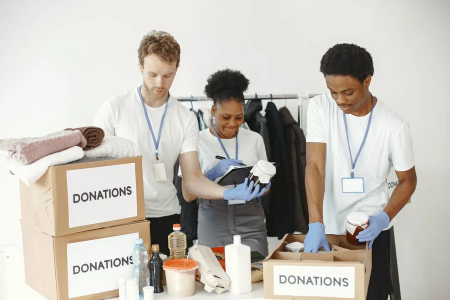 Three volunteers wearing ID badges sorting clothing and food into boxes marked DONATIONS at a community center