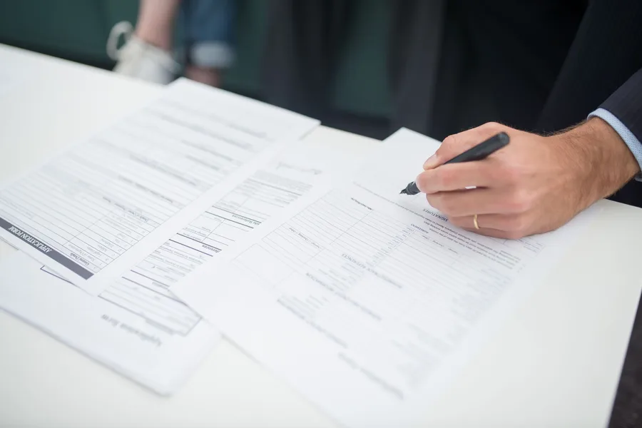A hand with a pen resting on printed application forms on a desk