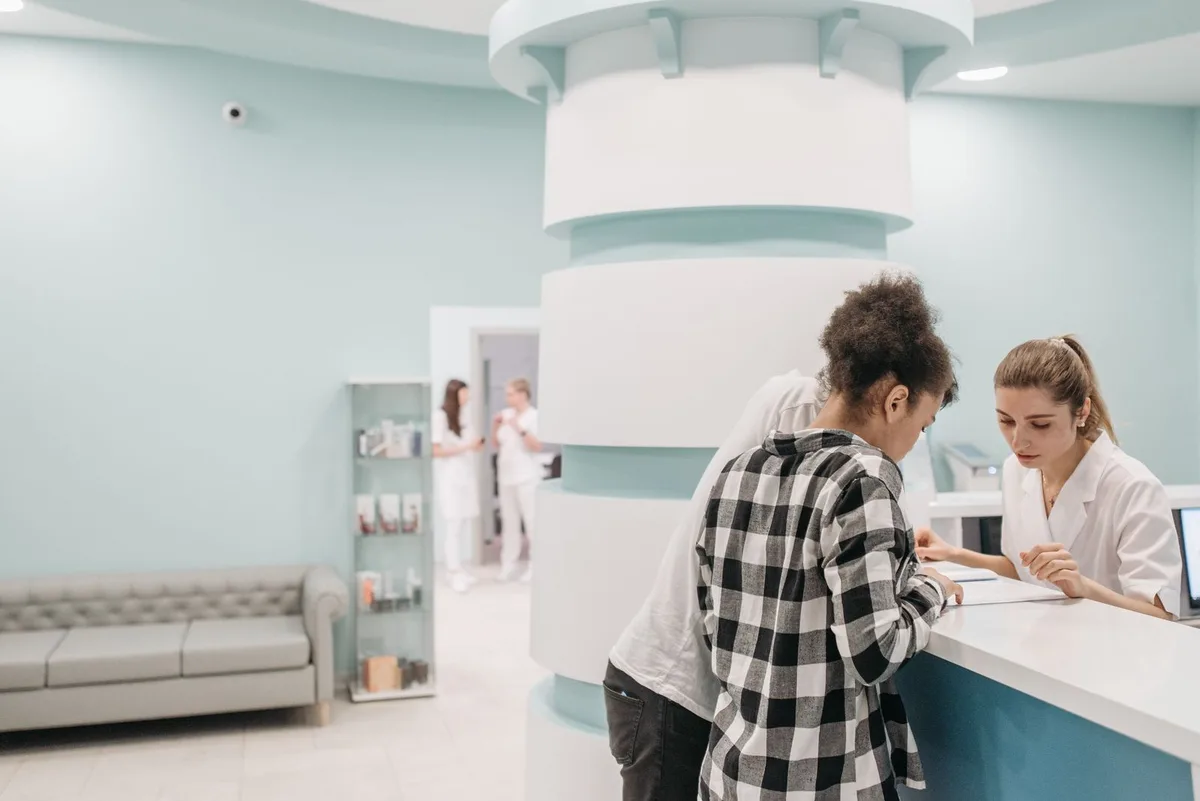 Two patients checking in at a clinic reception desk while a staff member helps with the paperwork