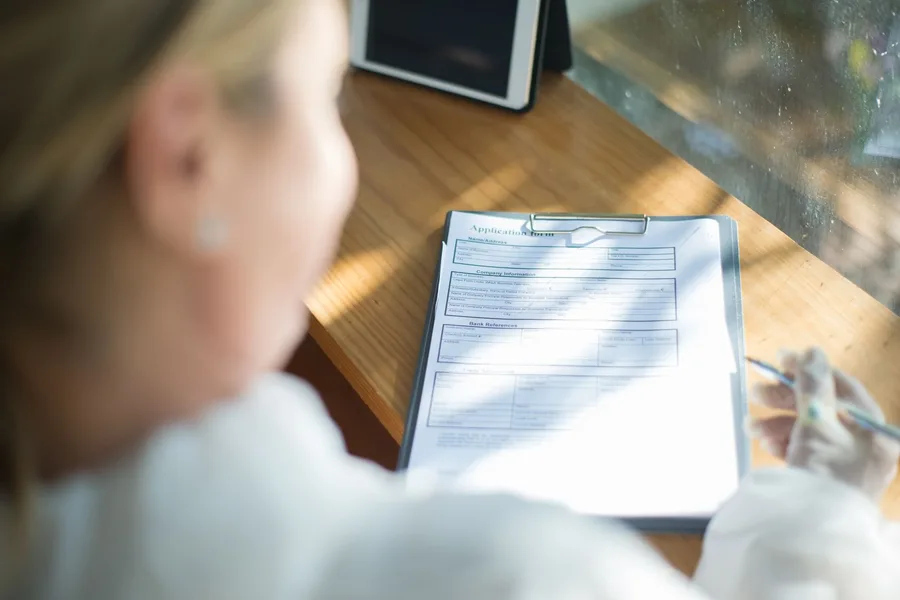A woman at a sunlit desk filling out a printed application form on a clipboard