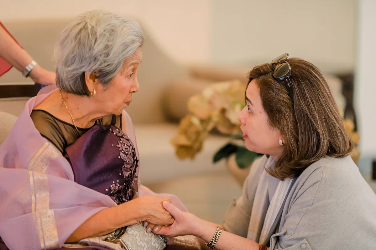 An older woman and her adult daughter sit close together at home, holding hands and talking quietly