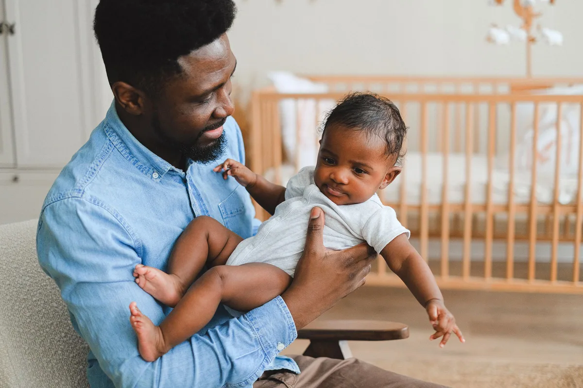 A father sitting in a nursery armchair holding his baby, with a wooden crib behind them