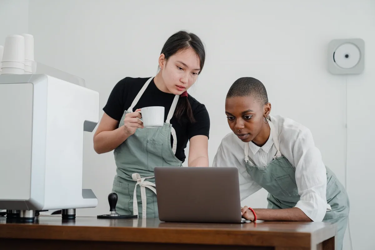 Two young cafe workers in aprons looking over a laptop together behind the counter