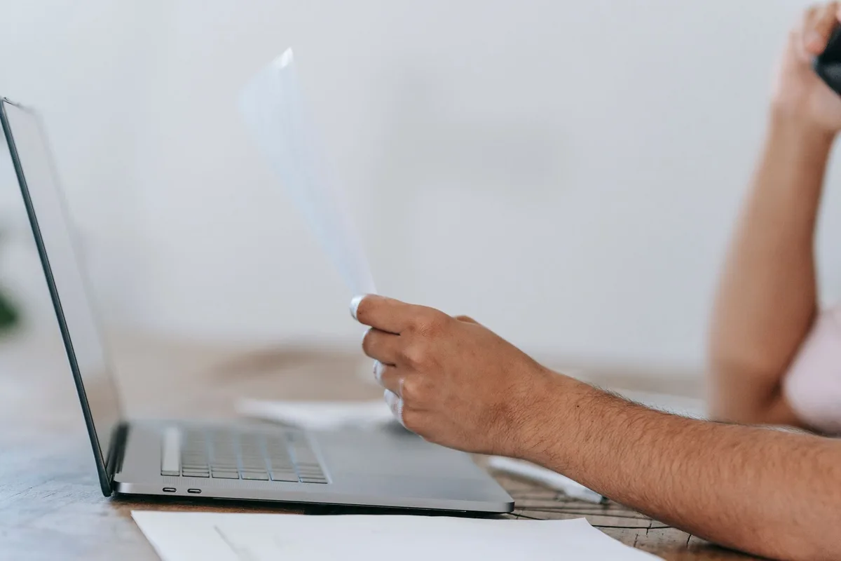 Hands holding a paper utility bill beside an open laptop while talking on the phone at home