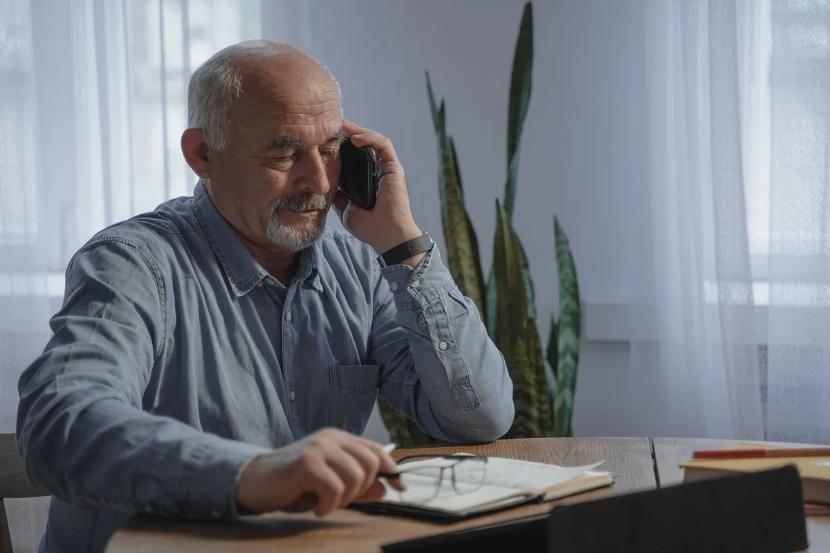 An older man sits at his dining table on a mobile phone with an open notebook, a pen and his glasses in front of him