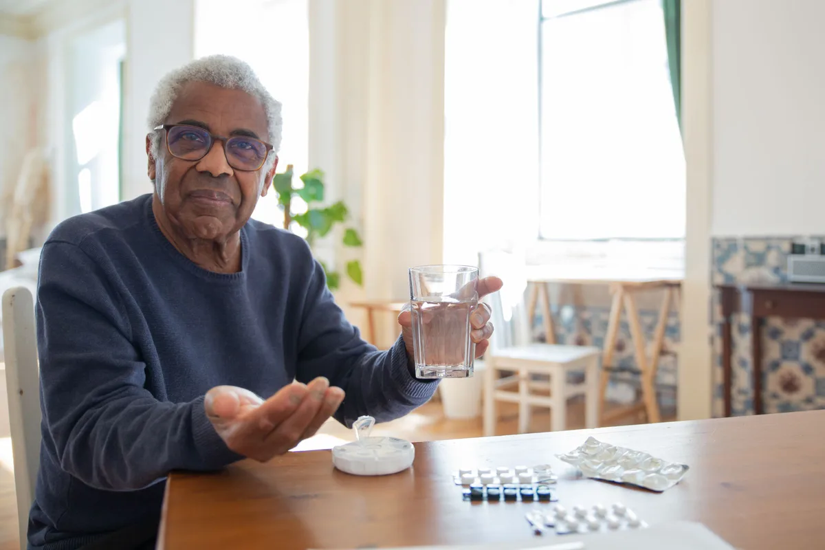 An older man sitting at a bright kitchen table with a glass of water and his morning pills laid out in front of him