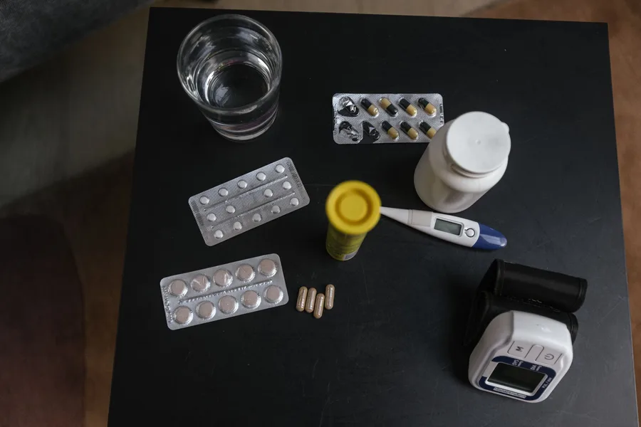 A side table holding blister packs of pills, a pill bottle, a thermometer, a blood pressure monitor and a glass of water