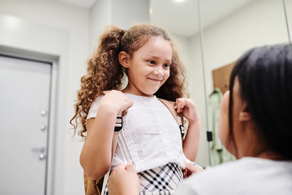 A mother helps her smiling daughter put on a school backpack in a hallway