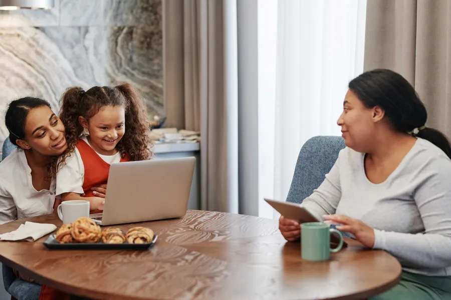 Two women and a young girl sit at a table with a laptop and a tablet