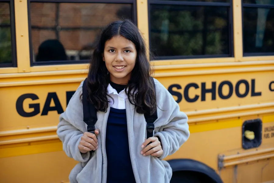 A smiling girl with a backpack stands in front of a yellow school bus