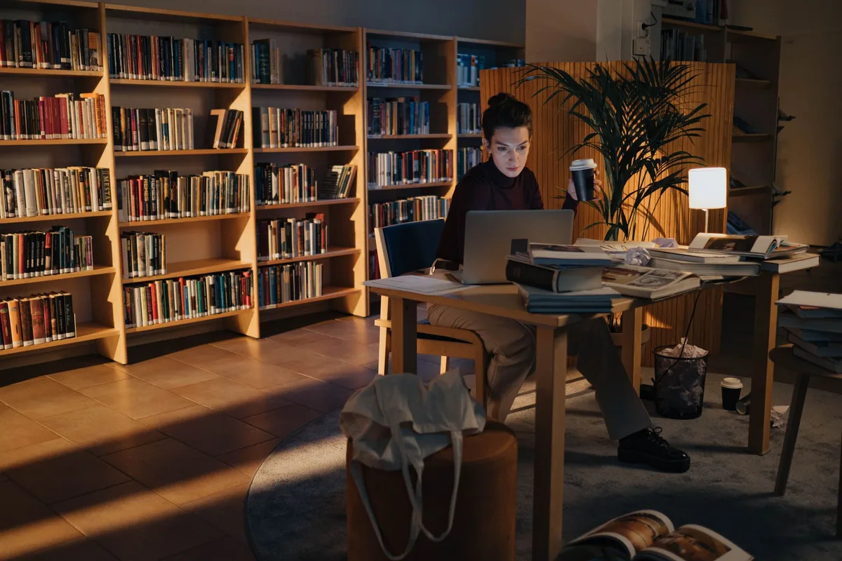 A student sitting at a desk among library shelves, working at a laptop in the evening