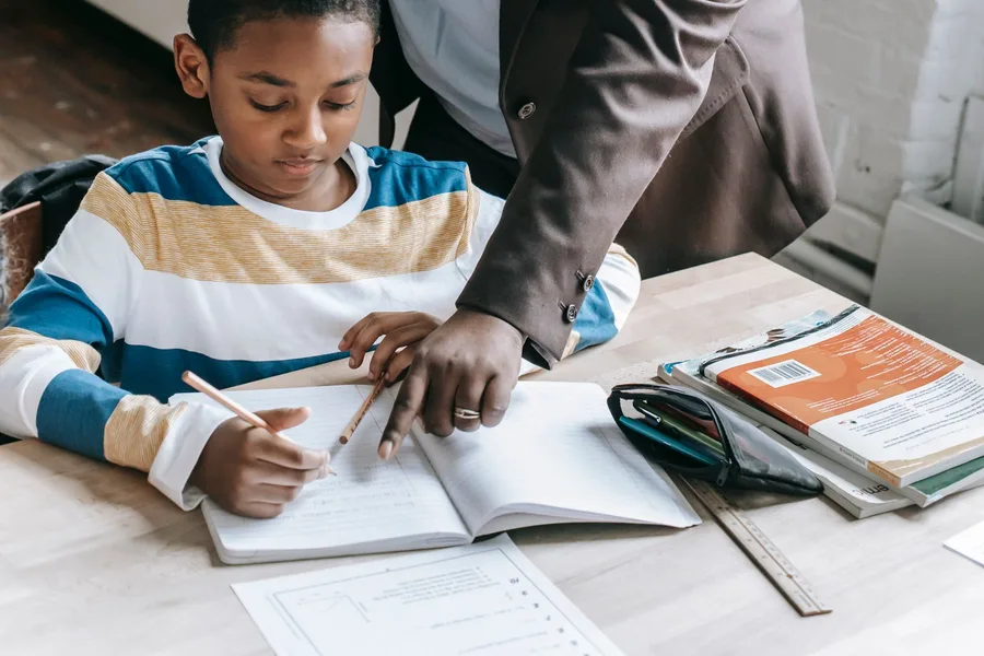 An adult guiding a child through schoolwork at a table, pointing at the open notebook