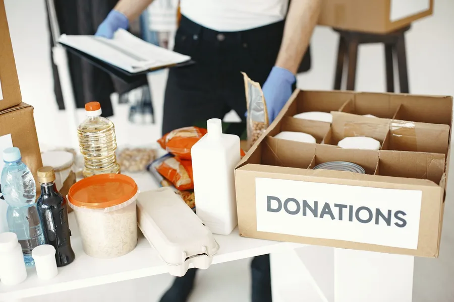 A donations box of canned goods beside rice, eggs, cooking oil and bottled water on a table, with a volunteer checking a clipboard behind it