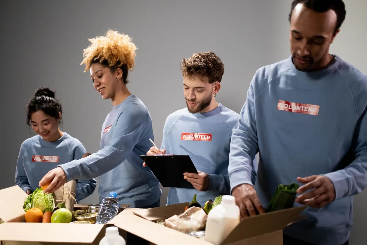 Four volunteers in blue VOLUNTEER sweatshirts packing cardboard boxes with lettuce, oranges, canned goods and milk at a food bank