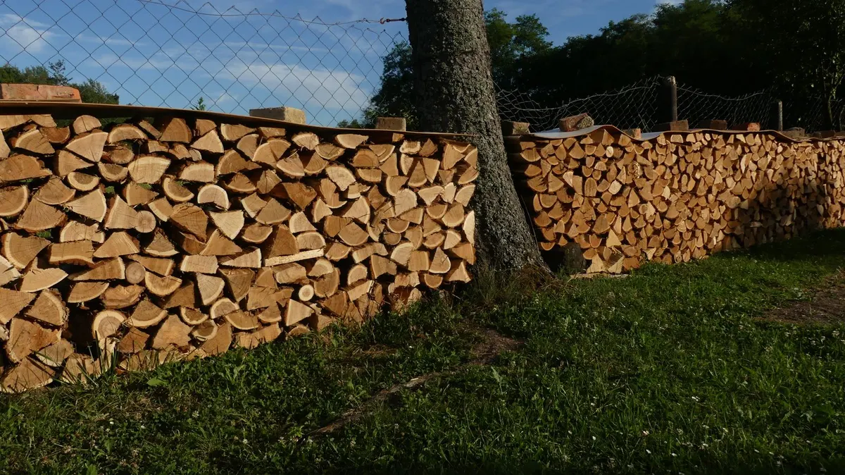 Split firewood stacked in two long rows along a wire fence, with boards weighted by bricks covering only the top of each stack