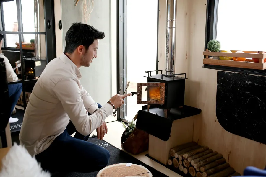 A man in a light shirt kneels beside a small black wood stove and opens its door, with cut logs stacked neatly underneath