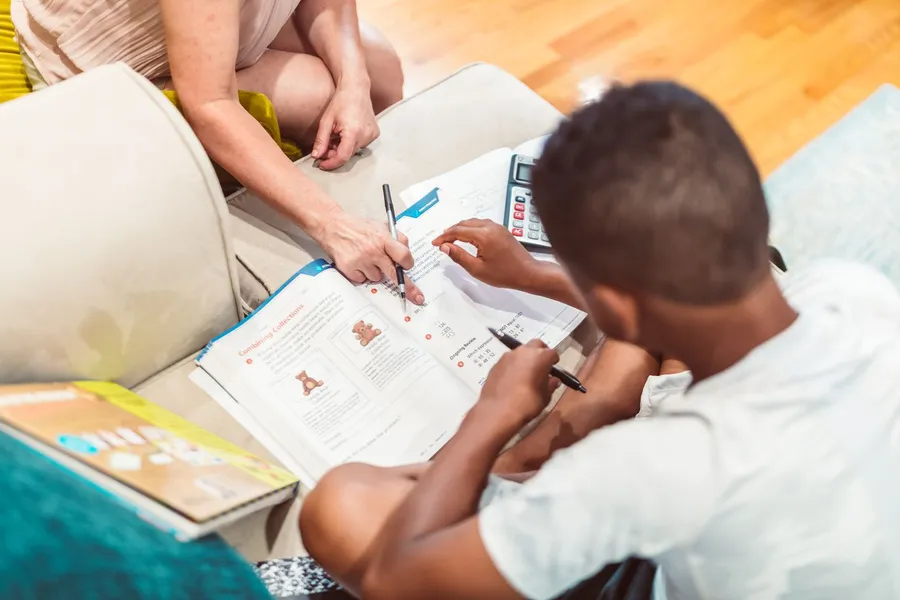 An adult points to a math worksheet while a boy holding a pen works through the problem