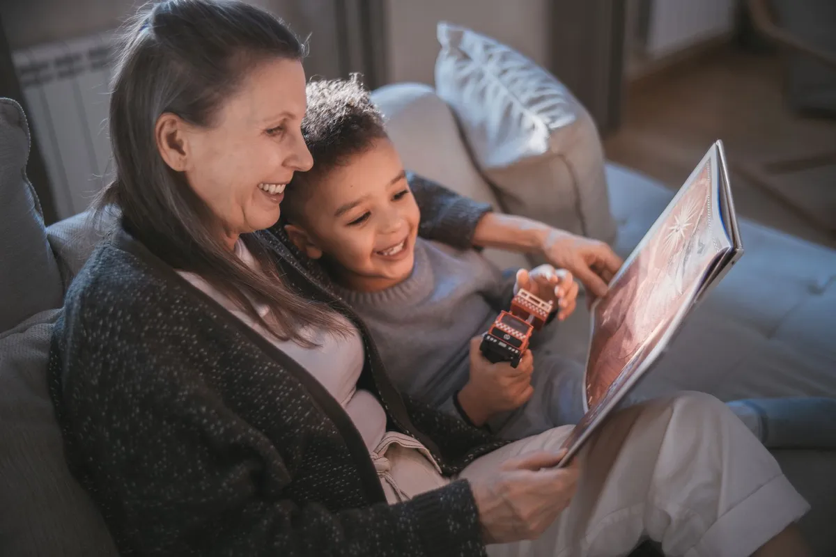 An older woman laughs as she reads a picture book with a young boy on a gray sofa
