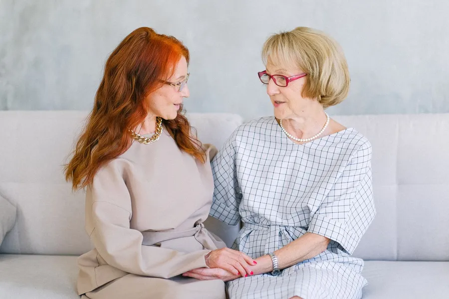 Two older women sit close together on a light sofa, holding hands and talking