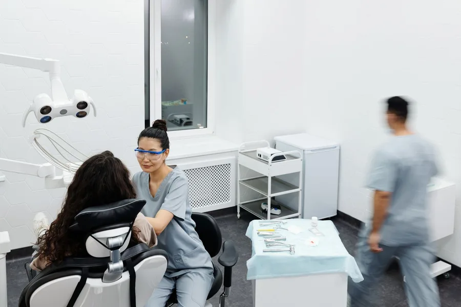 A dentist in scrubs sitting beside a patient in a dental chair in a bright treatment room, with an overhead light and a tray of instruments