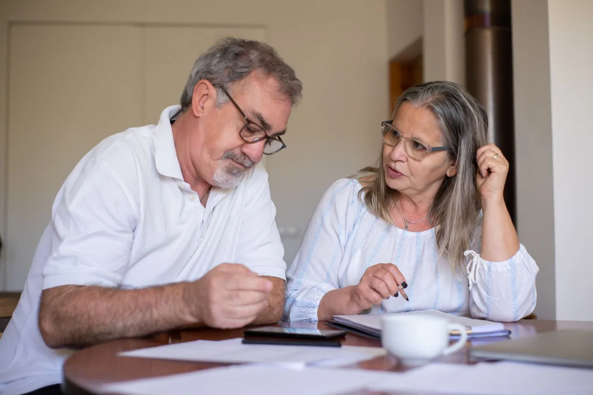 An older couple sitting at a dining table with printed papers, a notebook and a cup of coffee, talking something over