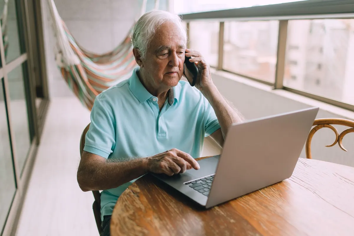 An older man with white hair sits at a wooden table on a sunny balcony, talking on his phone while looking at a laptop
