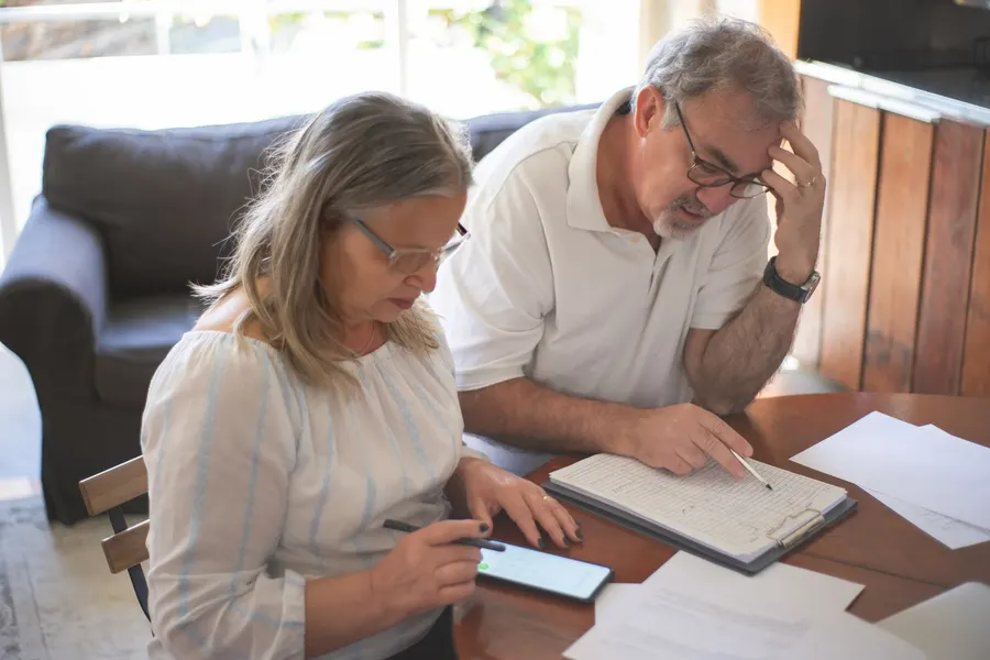 An older couple at a dining table go over a printed form on a clipboard together, with a phone beside them
