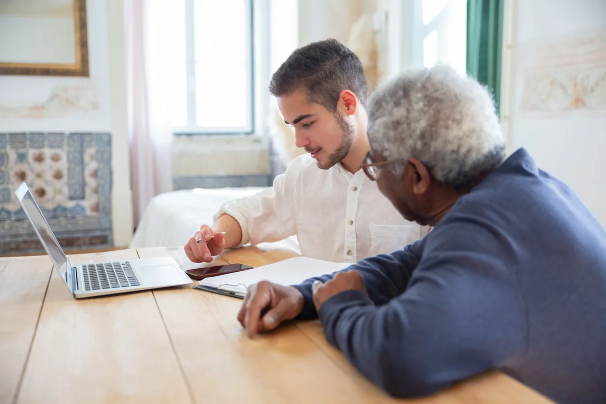 An older man and a younger volunteer sitting side by side at a wooden table, going over papers next to an open laptop