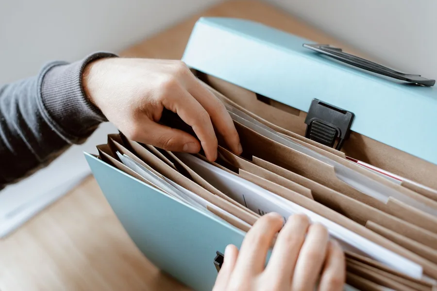 Two hands sorting paperwork into a pale blue expanding file folder on a wooden table