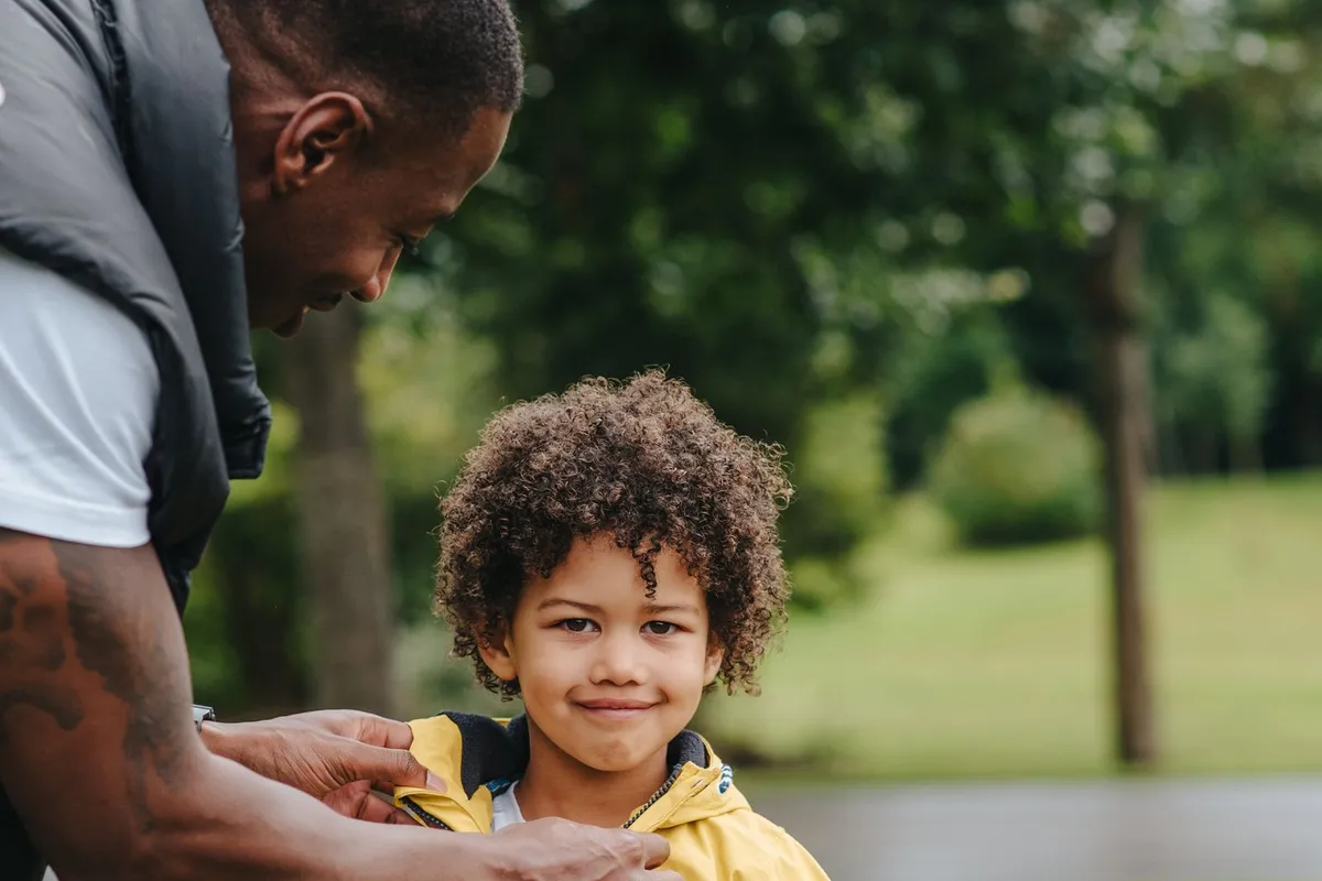 A father leans down to zip up his young son's yellow rain jacket in a park
