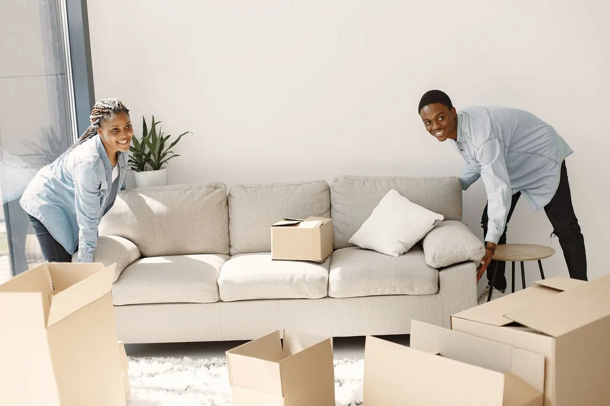 A smiling couple moves a light gray sofa into place in a sunny living room full of moving boxes