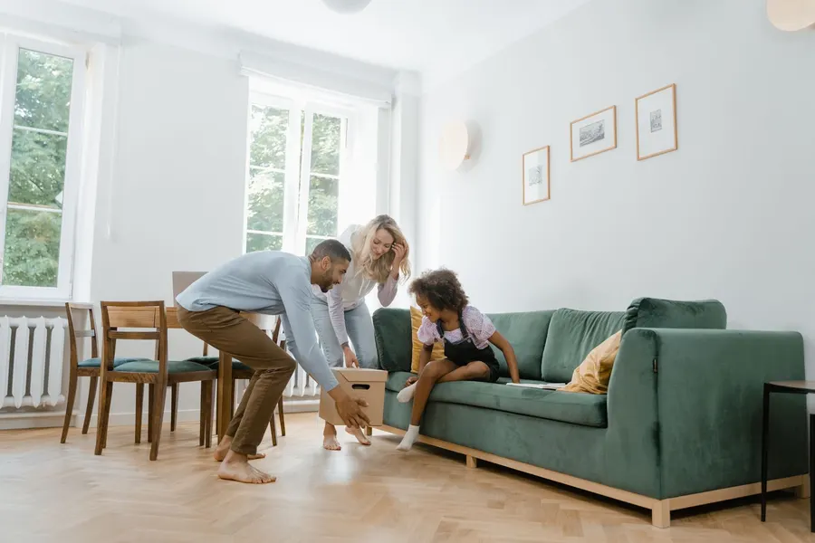 A father and mother lift a cardboard box as their young daughter sits smiling on a green sofa