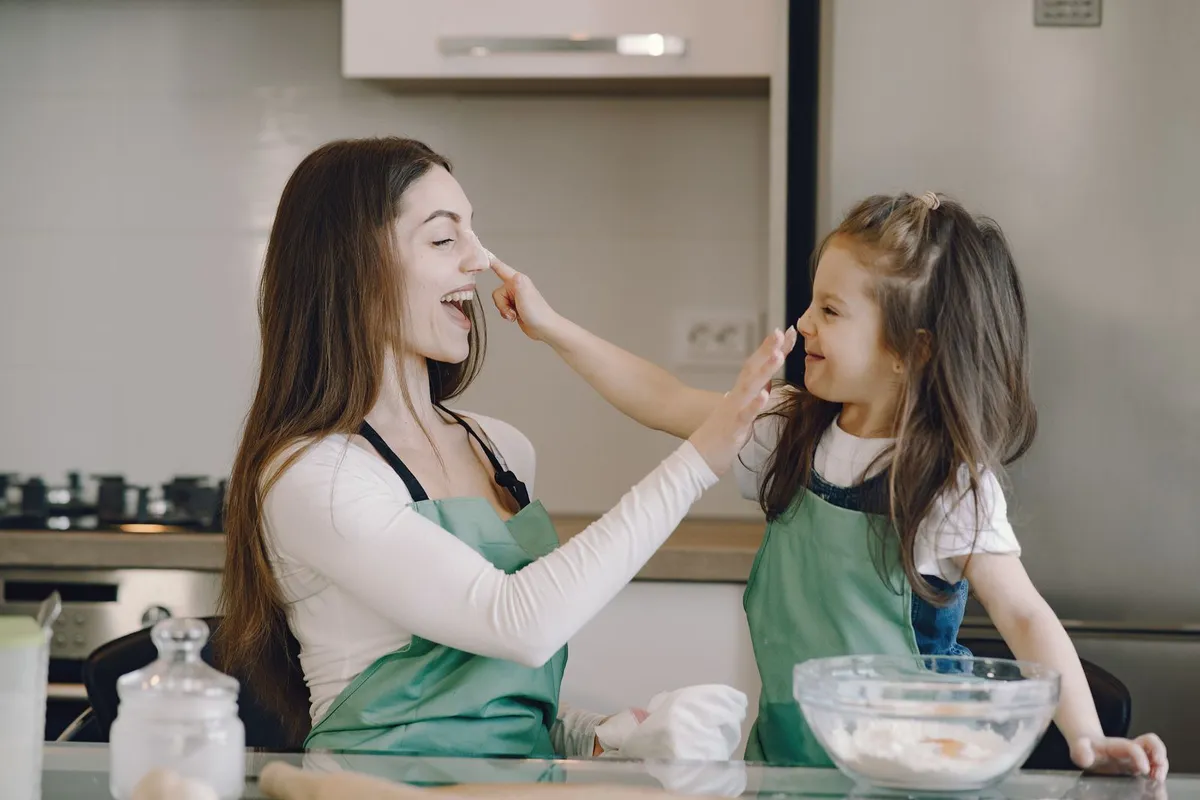 A mother and her young daughter laughing together while baking in their kitchen, dabs of flour on their noses