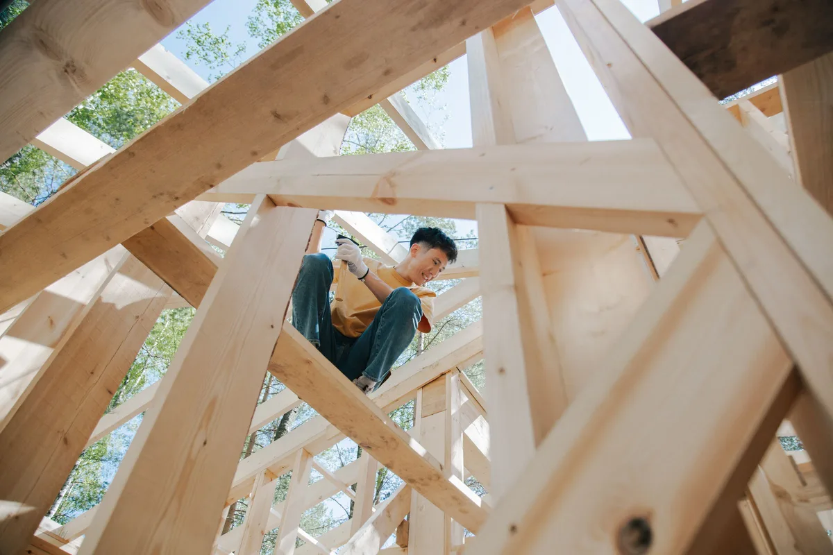 A person in work gloves smiling while fastening timber inside the wooden frame of a house under construction