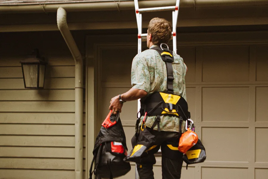A worker wearing a safety harness climbing a ladder toward the roofline of a single-story house