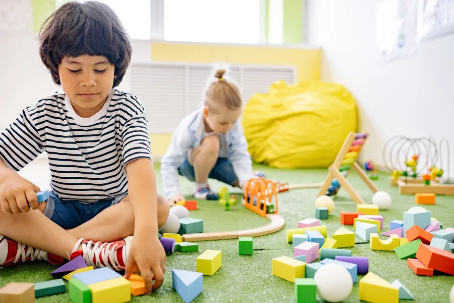 Two young children playing with wooden blocks on the floor of a bright classroom